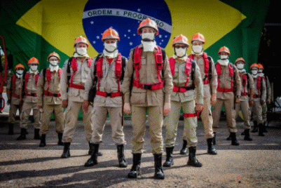 Equipe de bombeiros e brigadistas em fileira utilizando equipamentos de proteção individual (EPI) e capacetes laranjas com a bandeira do Brasil ao fundo.